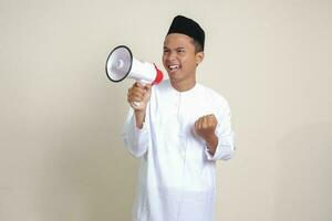 Portrait of attractive Asian muslim man in white shirt with skullcap speaking louder using megaphone, promoting product. Advertising concept. Isolated image on grey background photo