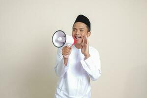 Portrait of attractive Asian muslim man in white shirt with skullcap speaking louder using megaphone, promoting product. Advertising concept. Isolated image on grey background photo