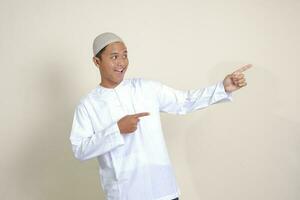 Portrait of attractive Asian muslim man in white shirt with skullcap showing product and pointing with his hand and finger to the side. Advertising concept. Isolated image on gray background photo