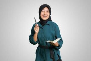 Portrait of excited Asian muslim woman with hijab writing on note book and pointing up with pen. Advertising concept. Isolated image on white background photo