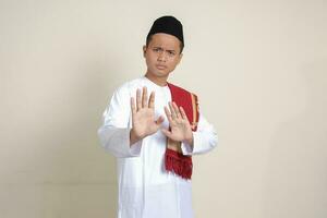 Portrait of attractive Asian muslim man in white shirt with skullcap forming a hand gesture to avoid something or bad things. Isolated image on grey background photo