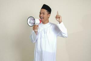 retrato de atractivo asiático musulmán hombre en blanco camisa con casquete Hablando mas fuerte utilizando megáfono, promoviendo producto. publicidad concepto. aislado imagen en gris antecedentes foto