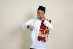 Portrait of attractive Asian muslim man in white shirt looking on wrist watch realizing that he is getting late for a meeting. Isolated image on gray background photo