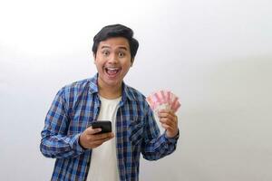 Portrait of excited Asian man in blue plaid shirt standing against white background, showing one hundred thousand rupiah while using mobile phone. Financial and savings concept. photo