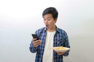 Portrait of happy Asian man in blue plaid shirt eating delicious instant noodles with chopsticks served on plate while using mobile phone. Isolated image on white background photo