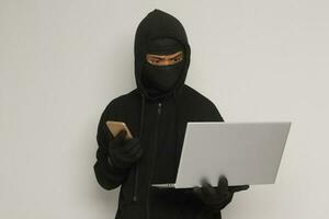 Portrait of mysterious man wearing black hoodie and mask doing hacking activity on laptop, hacker holding a mobile phone. Cyber security concept. Isolated image on gray background photo