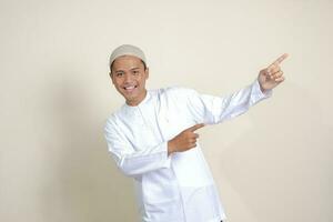 Portrait of attractive Asian muslim man in white shirt with skullcap showing product and pointing with his hand and finger to the side. Advertising concept. Isolated image on gray background photo