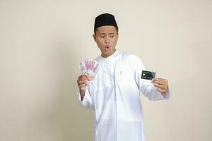 Portrait of attractive Asian muslim man in white shirt with skullcap holding one hundred thousand rupiah and presenting credit card. Isolated image on gray background photo