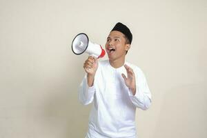 Portrait of attractive Asian muslim man in white shirt with skullcap speaking louder using megaphone, promoting product. Advertising concept. Isolated image on grey background photo