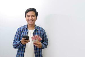 Portrait of excited Asian man in blue plaid shirt standing against white background, showing one hundred thousand rupiah while using mobile phone. Financial and savings concept. photo