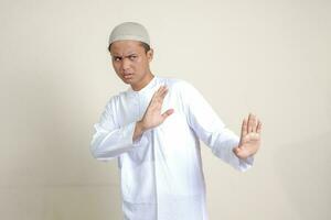 Portrait of attractive Asian muslim man in white shirt with skullcap forming a hand gesture to avoid something or bad things. Isolated image on grey background photo