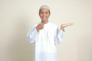 Portrait of attractive Asian muslim man in white shirt with skullcap showing product and pointing with his hand and finger to the side. Advertising concept. Isolated image on gray background photo