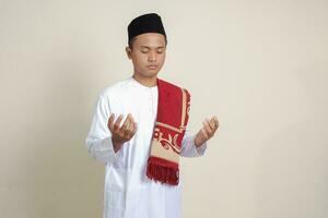 Portrait of attractive Asian muslim man in white shirt with skullcap praying earnestly with his hands raised. Isolated image on gray background photo