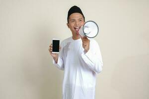 Portrait of attractive Asian muslim man in white shirt showing and presenting blank screen mobile phone while speaking loudly with megaphone. Advertising concept. Isolated image on gray photo