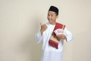 Portrait of attractive Asian muslim man in white shirt showing one hundred thousand rupiah while pointing to the side. Financial and savings concept. Isolated image on gray background photo