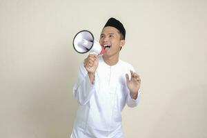 Portrait of attractive Asian muslim man in white shirt with skullcap speaking louder using megaphone, promoting product. Advertising concept. Isolated image on grey background photo