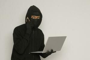 Portrait of mysterious man wearing black hoodie and mask doing hacking activity on laptop, hacker holding a mobile phone. Cyber security concept. Isolated image on gray background photo