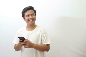 Portrait of shocked Asian man in white t-shirt standing against white background, holding a mobile phone while making surprise hand gesture. Wow face expression. Advertising concept. photo