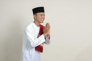 Portrait of attractive Asian muslim man in white shirt with skullcap showing apologize and welcome hand gesture. Isolated image on gray background photo