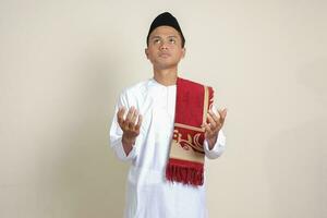 Portrait of attractive Asian muslim man in white shirt with skullcap praying earnestly with his hands raised. Isolated image on gray background photo