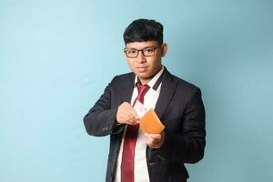 Portrait of young Asian business man in casual suit holding leather wallet and pulling out thousand rupiahs while looking at camera. Isolated image on blue background photo