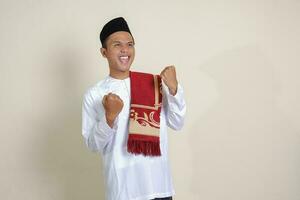 Portrait of attractive Asian muslim man in white shirt raising his fist, celebrating success. Isolated image on gray background photo