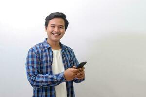 Portrait of excited Asian man in blue plaid shirt looking at camera and holding mobile phone. Isolated image on white background photo