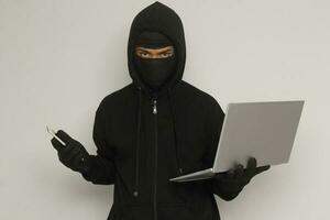 Portrait of mysterious man wearing black hoodie and mask doing hacking activity on laptop, hacker holding a personal computer. Cyber security concept. Isolated image on gray background photo