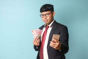 Portrait of young Asian business man in casual suit showing phone and holding thousand rupiahs looking at camera while smiling. Isolated image on blue background photo