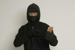 Portrait of mysterious man wearing black hoodie and mask doing hacking activity on mobile phone, hacker holding a smartphone. Isolated image on gray background photo