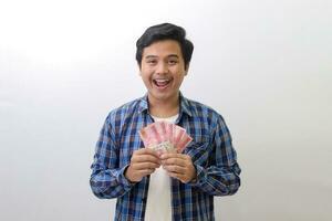 Portrait of excited Asian man in blue plaid shirt standing against white background, showing one hundred thousand rupiah while pointing to the side. Financial and savings concept. photo