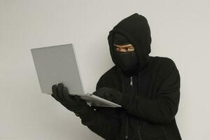 Portrait of mysterious man wearing black hoodie and mask doing hacking activity on laptop, hacker holding a personal computer. Cyber security concept. Isolated image on gray background photo