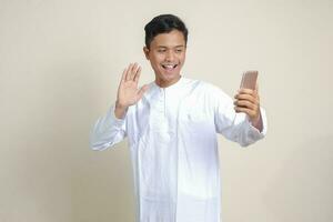 retrato de atractivo asiático musulmán hombre en blanco camisa tomando imagen de él mismo o autofoto, diciendo Hola y ondulación su mano durante vídeo llamar. aislado imagen en gris antecedentes foto