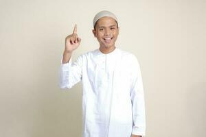 Portrait of attractive Asian muslim man in white shirt pointing up his forefinger and reminding something to do. Advertising concept. Isolated image on gray background photo