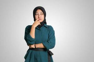 Portrait of confused Asian muslim woman with hijab standing against white background, looking up and thinking about question with hand on chin photo