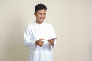 Portrait of attractive Asian muslim man in white shirt holding mobile phone with smiling expression on face. Advertising concept. Isolated image on gray photo