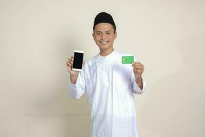 Portrait of attractive Asian muslim man in white shirt with skullcap holding a mobile phone and presenting credit card. Isolated image on gray background photo