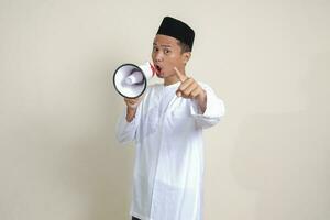 Portrait of attractive Asian muslim man in white shirt with skullcap speaking louder using megaphone, promoting product. Advertising concept. Isolated image on grey background photo