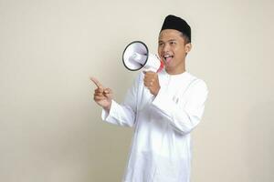 Portrait of attractive Asian muslim man in white shirt with skullcap speaking louder using megaphone, promoting product. Advertising concept. Isolated image on grey background photo