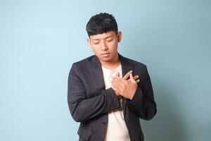 Portrait of young Asian business man in casual suit heart broken holding chest with both hands. Isolated image on blue background photo