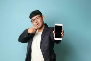 Portrait of young Asian business man in casual suit showing at camera while pointing at phone looking at camera. Isolated image on blue background photo