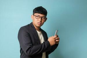 Portrait of young Asian business man in casual suit looking surprised at the camera while holding phone. Isolated image on blue background photo