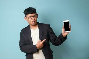 Portrait of young Asian business man in casual suit pointing at phone with confused expressions. Isolated image on blue background photo