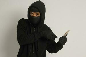 Portrait of mysterious man wearing black hoodie and mask doing hacking activity on mobile phone, hacker holding a smartphone. Isolated image on gray background photo