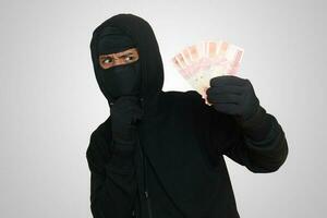 Portrait of mysterious man wearing black hoodie and mask stealing one hundred thousand rupiah successfully from victim. Isolated image on gray background photo