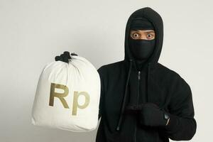 Mysterious robber thief man wearing black hoodie and mask carrying a bag full of rupiah money. Isolated image on gray background photo