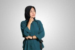 Portrait of confused Asian muslim woman with hijab standing against white background, looking up and thinking about question with hand on chin photo