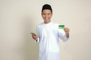 Portrait of attractive Asian muslim man in white shirt with skullcap holding a mobile phone and presenting credit card. Isolated image on gray background photo