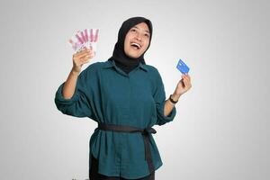 Portrait of cheerful Asian muslim woman with hijab, showing one hundred thousand rupiah while holding a credit card. Financial and savings concept. Isolated image on white background photo