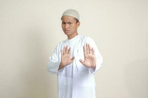 Portrait of attractive Asian muslim man in white shirt with skullcap forming a hand gesture to avoid something or bad things. Isolated image on grey background photo